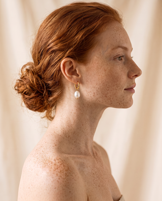 Pearl Drop Huggies in gold worn on the ear by a model with red hair, side profile