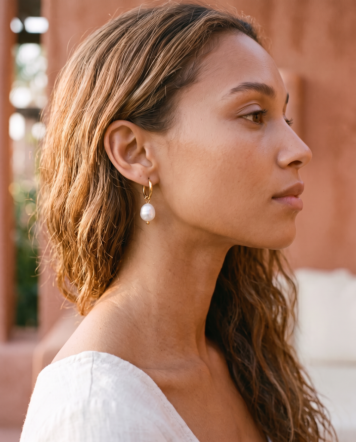 Pearl Drop Huggies in gold worn on the ear by a model with wavy hair, side profile